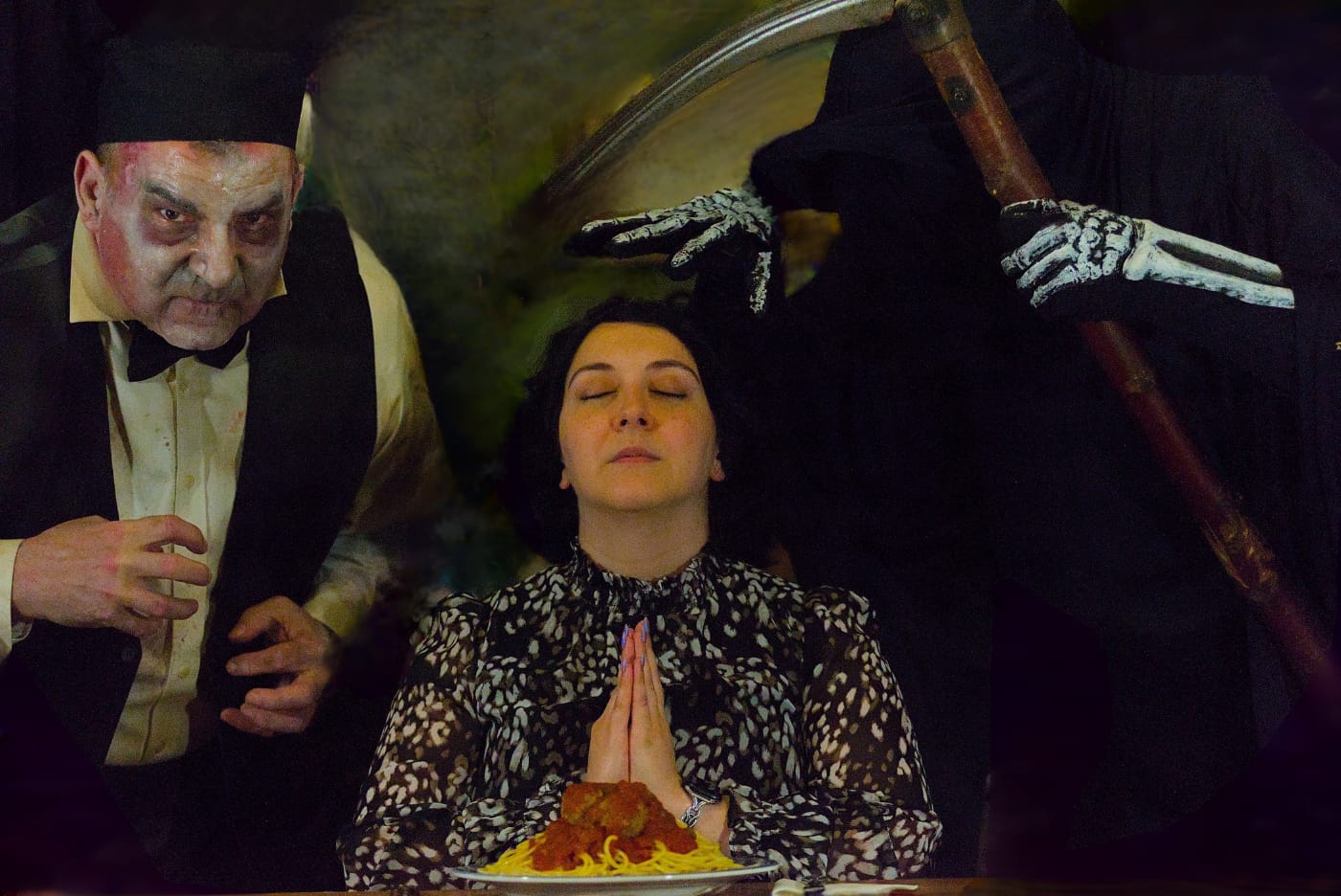 A diner sits with hands pressed together while a performer in zombie make-up and a hooded grim reaper with a scythe loom on either side